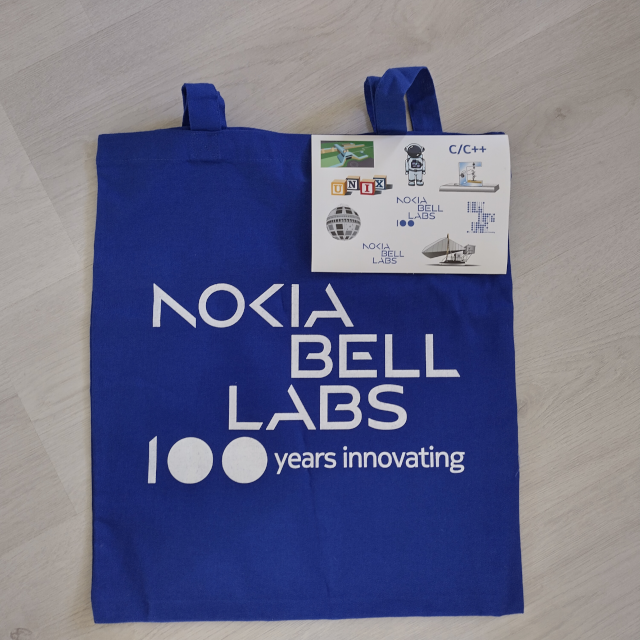 A stylish reusable shopping bag in Nokia blue color with white text saying "Nokia Bell Labs - 100 years innovating. There is also a sticker sheet depicting various things: building blocks with the letters UNIX, the Telstar 1 satellite, the Bell Labs HQ in Murray Hill, an astronaut, the first transistor, the Holmdel Horn Antenna that discovered the cosmic microwave radiation, and some logos.