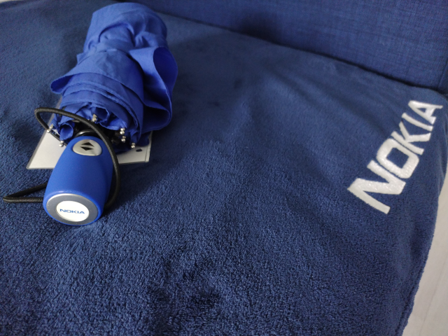A plain blue folding umbrella lies closed on a darker blue blanket. The handle of the umbrella features the old Nokia logo. The same logo is also embroidered in white onto the blanket.