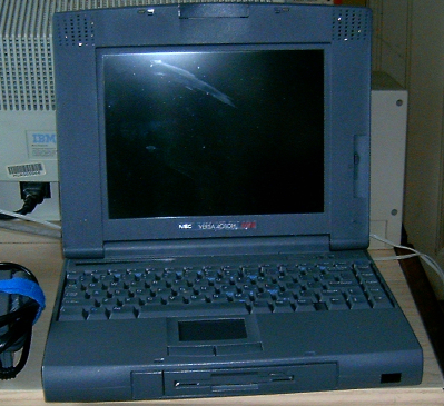 A 14-inch gray notebook from the nineties. The lid is open, but the machine is turned off. It has a built-in floppy drive at the front and an infrared port. Speakers are built into the display bezel at the top. There is also a manual brightness control slider below one of the speakers. In the background, the back of a 17-inch IBM CRT monitor is visible.