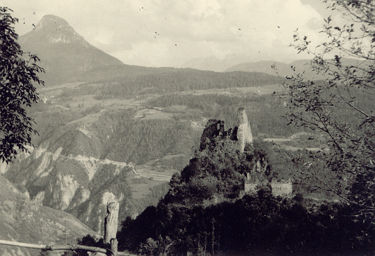 This is a high-quality scan of a photograph from from the 1920s. It was black and white, but it got a sepia tone. The top of a weak wooden fence is the closest thing. This is between the photographer and a steep ravine. On the next mountaintop there is a ruined castle tower partially covered by trees. Behind the ruin, far in a deep valley, there is a road going through a forest. High mountains can be seen on the horizon.