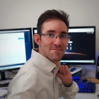 It's me sitting at a desk, turning away from the two displays in the background to look at the camera. I'm wearing a white shirt.